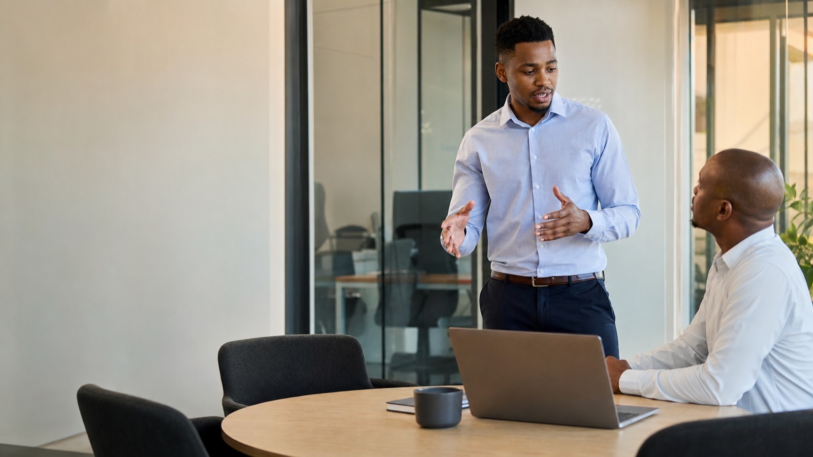 A Black South African businessman in a collared shirt gesturing while meeting with a client at a table in a small consulting office.