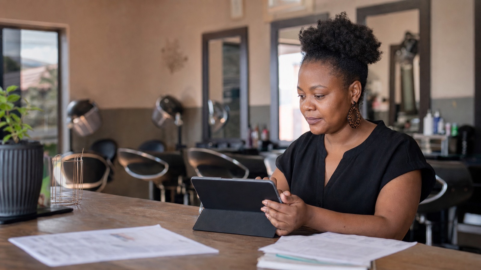 Black South African woman at a salon reception counter with a tablet and documents, styling chairs and mirrors behind her.