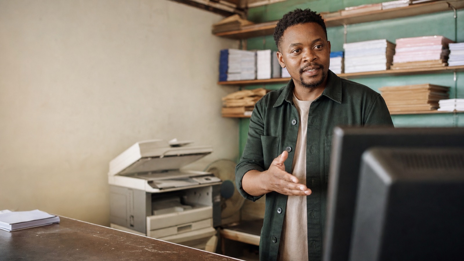 A man in a township copy shop gesturing toward a blurred computer screen, with a photocopier and paper supplies behind him.