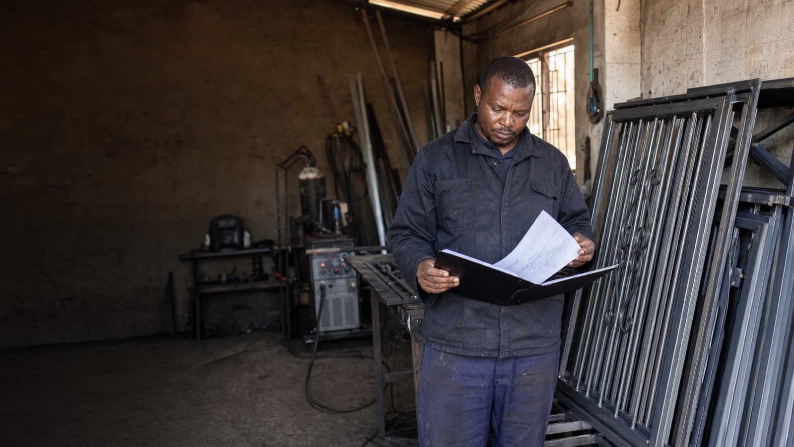 A Black South African welding business owner stands among welding equipment and metal products in his workshop, holding open a folder of documents.