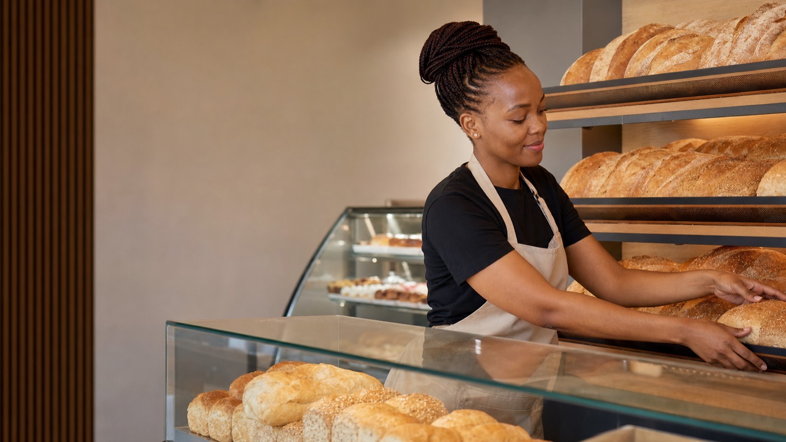 A young Black woman arranging fresh bread on display shelves inside a modern bakery franchise shop.