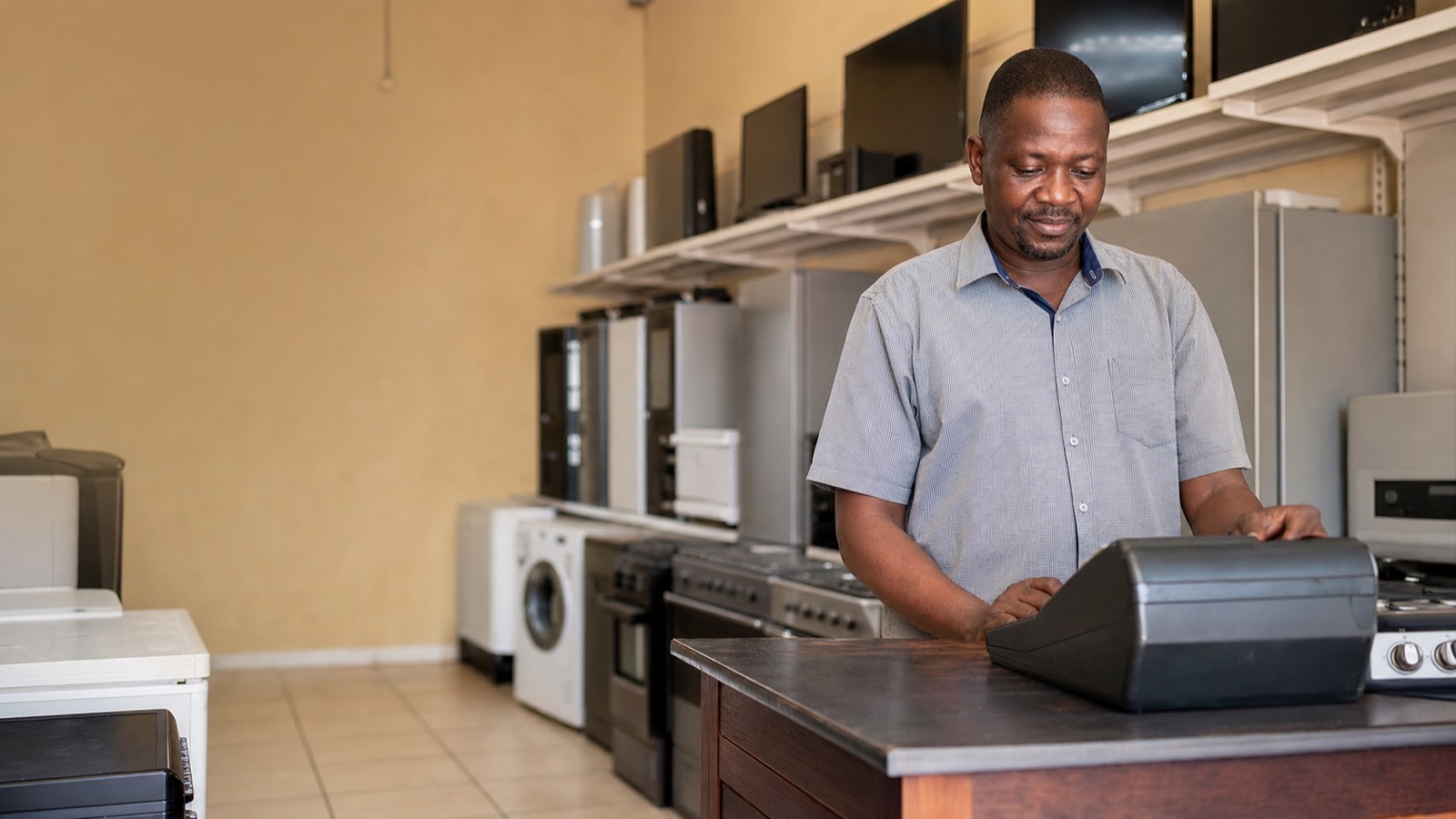 Black South African man processing a sale at the till counter of his township appliance shop with washing machines and TVs on shelves behind him.