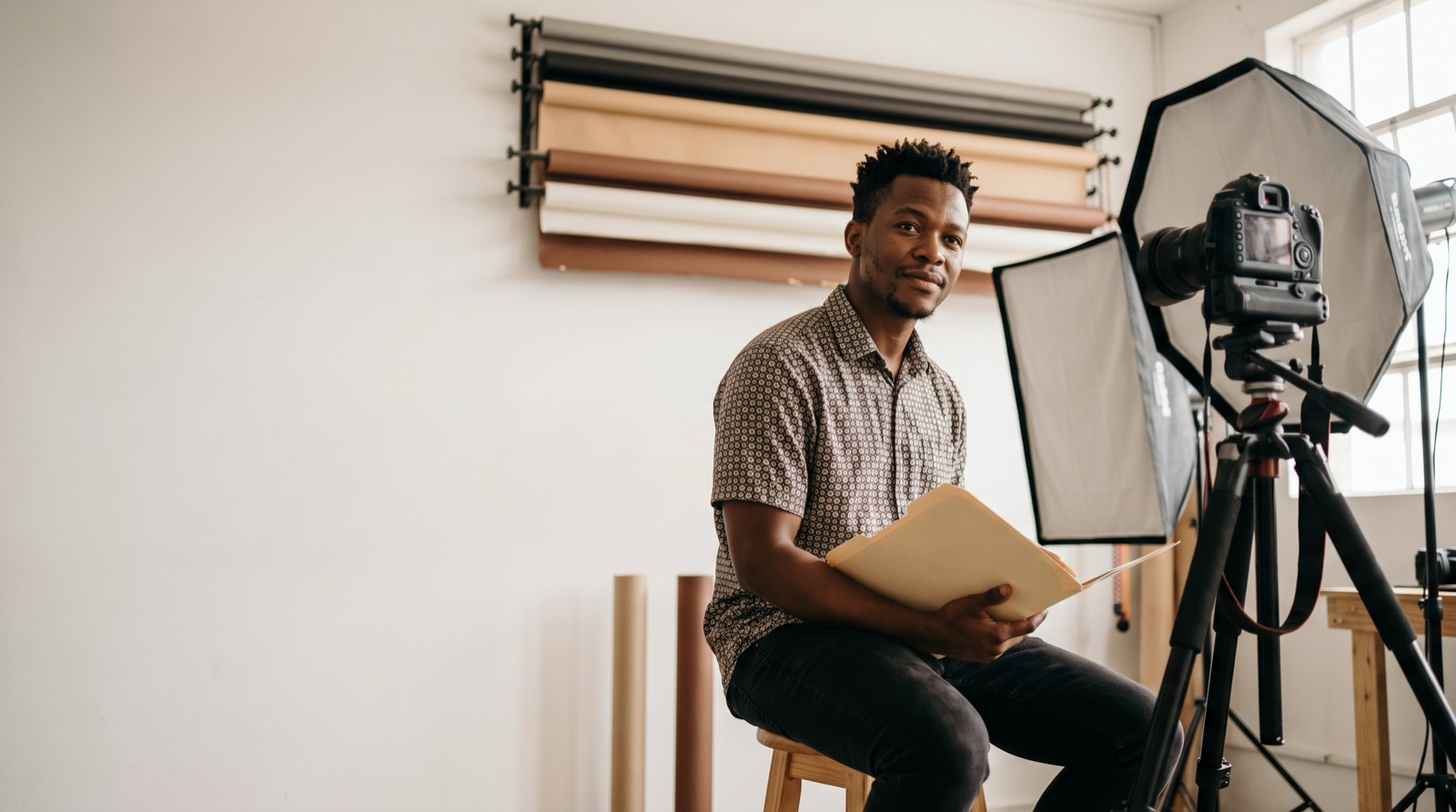 A young Black man sitting in a photography studio holding a folder, with camera equipment and backdrops behind him.