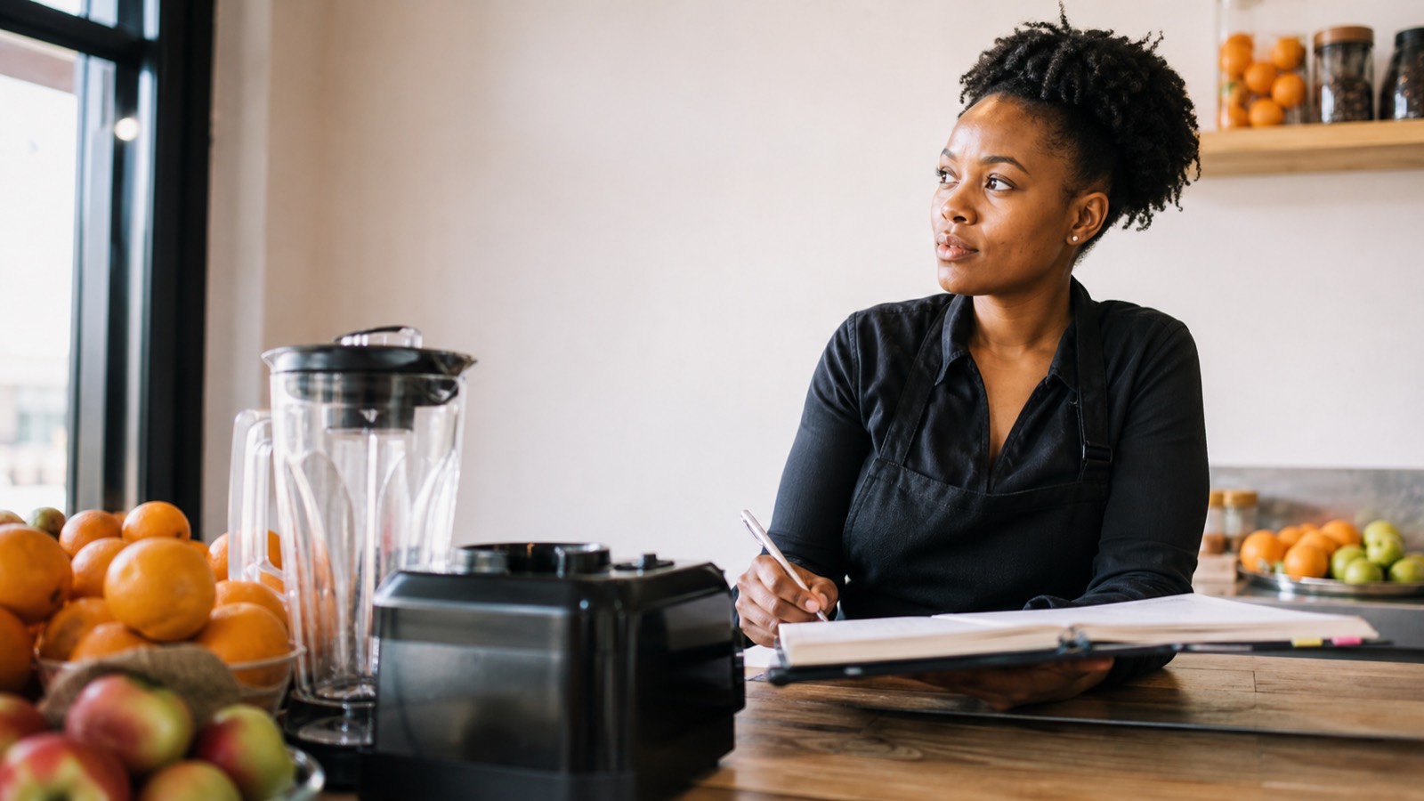 Young Black woman behind a juice bar counter with fresh fruit and a blender, reviewing business plan materials.
